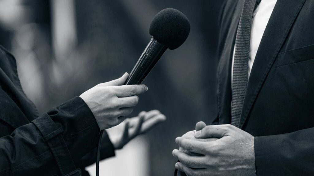 black and white closeup of an interviewer with microphone pointed at man in suit and tie